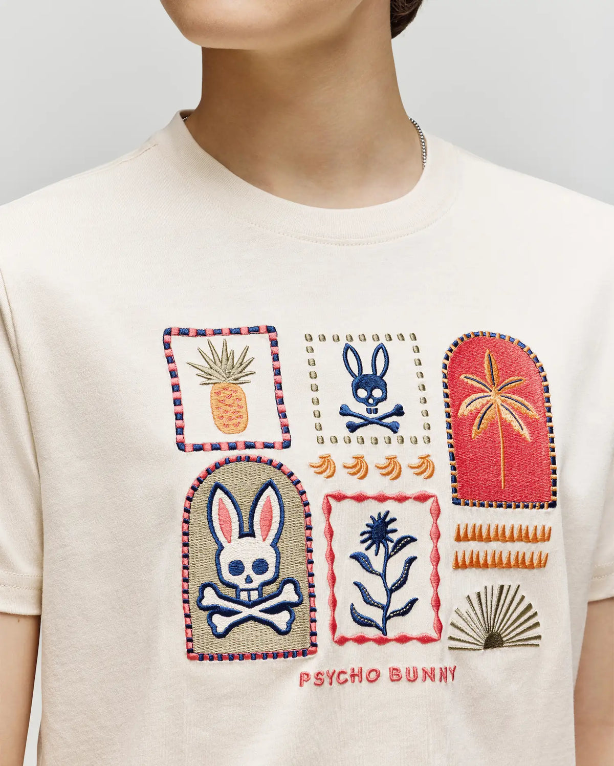 A child wears the Psycho Bunny KIDS SUNDEW GRAPHIC TEE (KT0200092), a cream Pima cotton shirt with vibrant embroidery—bunnies, skulls, plants, and "PSYCHO BUNNY" on the front. Only their lower face and upper torso are shown—ocean view style.