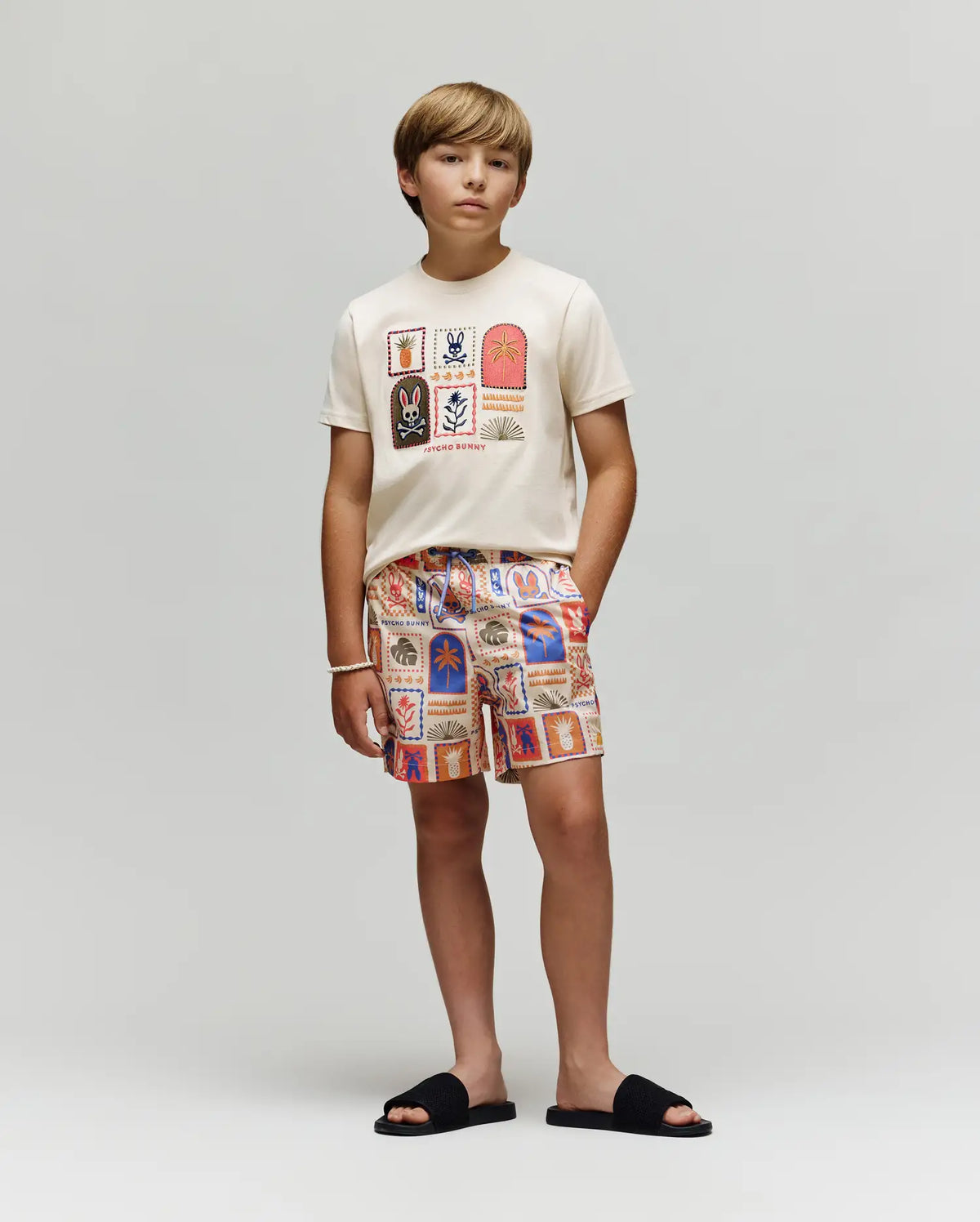 A young boy stands against a plain background, wearing the Psycho Bunny KIDS SUNDEW GRAPHIC TEE (KT0200092) in cream with colorful Beach Bunny prints, matching patterned shorts, black slide sandals, and a white beaded bracelet.