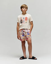 A young boy stands against a plain background, wearing the Psycho Bunny KIDS SUNDEW GRAPHIC TEE (KT0200092) in cream with colorful Beach Bunny prints, matching patterned shorts, black slide sandals, and a white beaded bracelet.