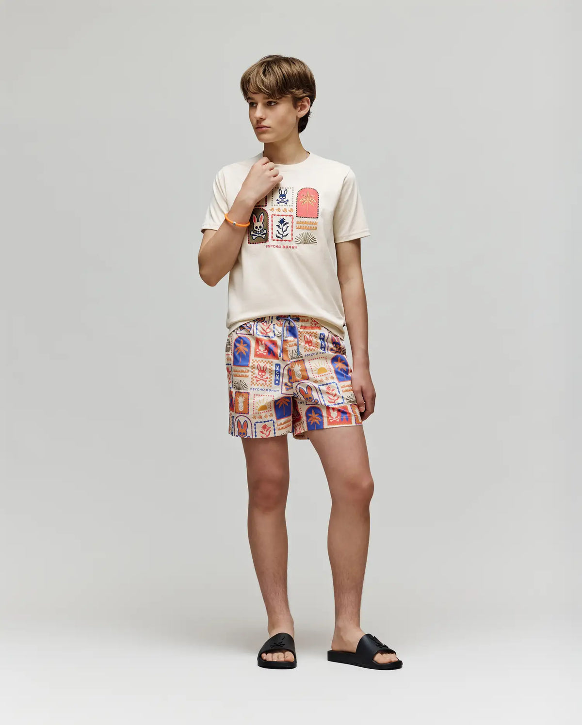 A young person stands against a plain background wearing the cream-colored Psycho Bunny KIDS SUNDEW GRAPHIC TEE (KT0200092), Beach Bunny print shorts, black slide sandals, and gazes thoughtfully to the side with a hand on their chin.