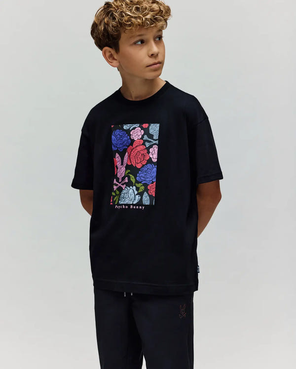 A young boy with curly hair wears the Psycho Bunny KIDS ROWEN OVERSIZED TEE (KT0200093) with a vibrant floral graphic, standing with hands behind his back and looking to the side against a plain light background.