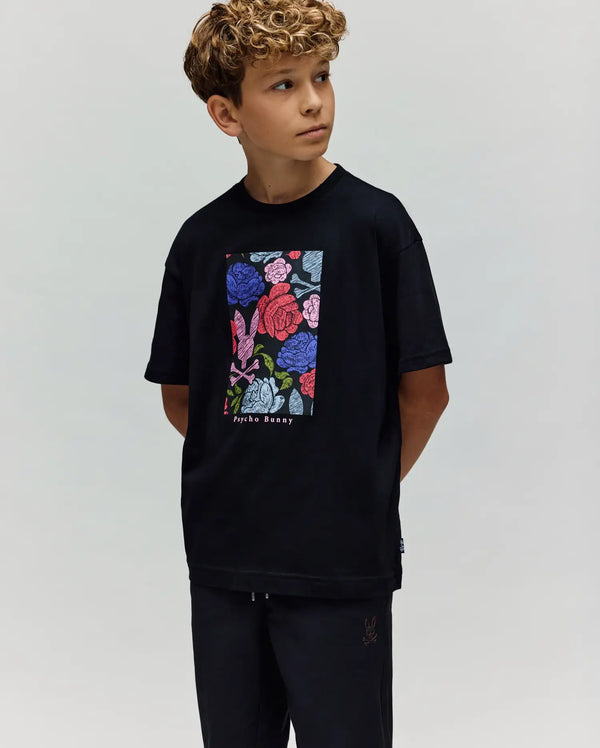 A young boy with curly hair wears the Psycho Bunny KIDS ROWEN OVERSIZED TEE (KT0200093) with a vibrant floral graphic, standing with hands behind his back and looking to the side against a plain light background.