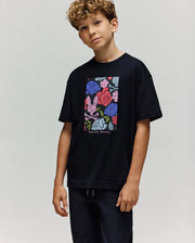 A young boy with curly hair wears the Psycho Bunny KIDS ROWEN OVERSIZED TEE (KT0200093), featuring a colorful floral graphic and "Psycho Bunny" text on the front. He stands against a plain, light background.