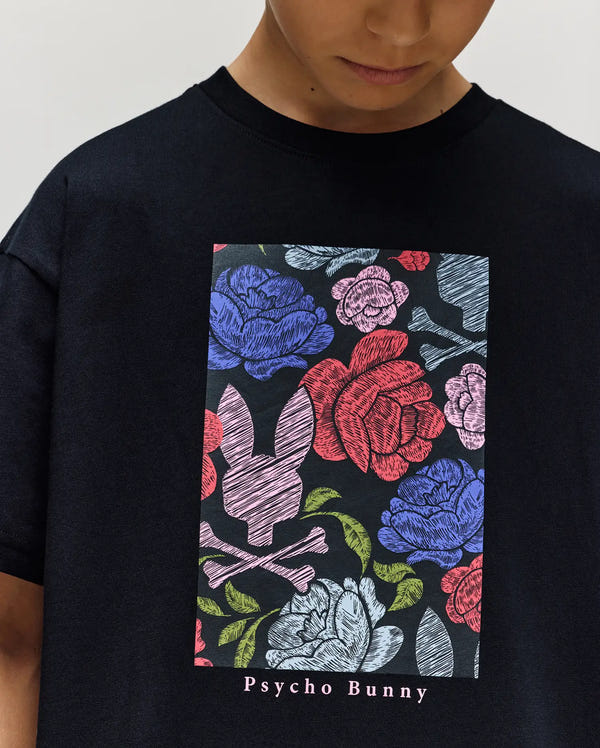 A child wears the Psycho Bunny KIDS ROWEN OVERSIZED TEE (KT0200093), a black relaxed fit t-shirt with a colorful rose and leaf graphic, plus a bunny head and crossbones. "Psycho Bunny" text appears below the floral design.