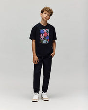 A boy with curly hair stands against a plain background, wearing the Psycho Bunny KIDS ROWEN OVERSIZED TEE (KT0200093), black pants, and white sneakers. He has one hand by his side and a relaxed posture.