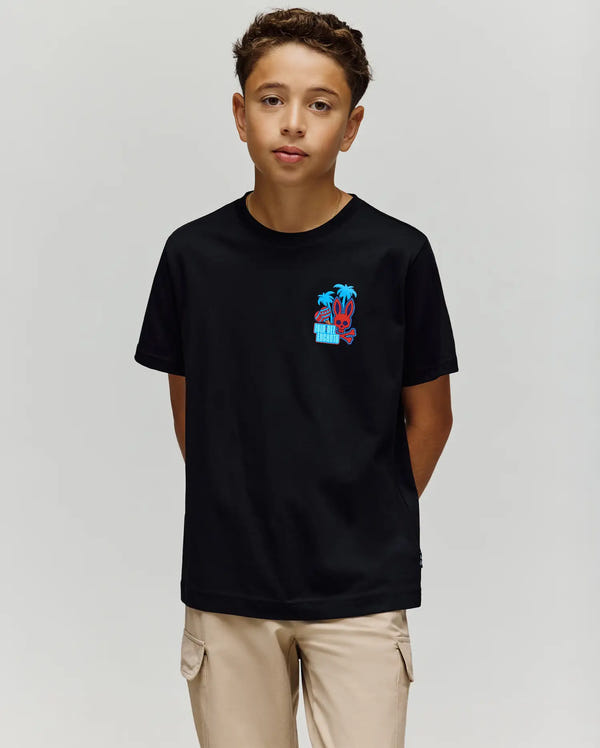 A boy stands against a plain light background wearing the Psycho Bunny KIDS PUERTO RICO TEE - KT0200133, a black Pima cotton t-shirt with a small colorful bunny graphic on the left chest, and light beige pants, hands in his pockets.