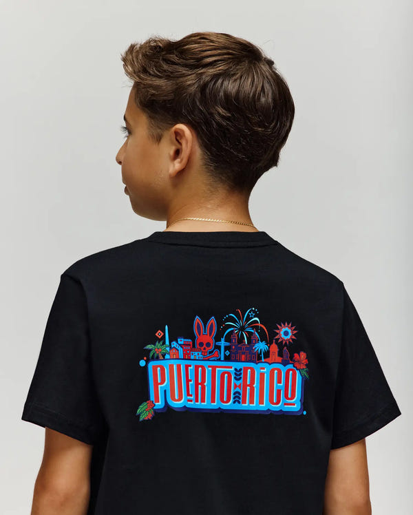 A boy is seen from the back wearing the Psycho Bunny KIDS PUERTO RICO TEE (KT0200133), a black Pima cotton shirt with a colorful Puerto Rico graphic featuring landmarks, palm trees, fireworks, and bold red, blue, and white lettering.