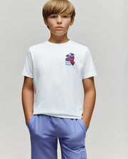 A young boy with blond hair stands against a plain background, wearing the Psycho Bunny KIDS PUERTO RICO TEE (KT0200133), featuring a colorful bunny graphic, and light blue shorts. His hands are in his pockets and he has a neutral expression.