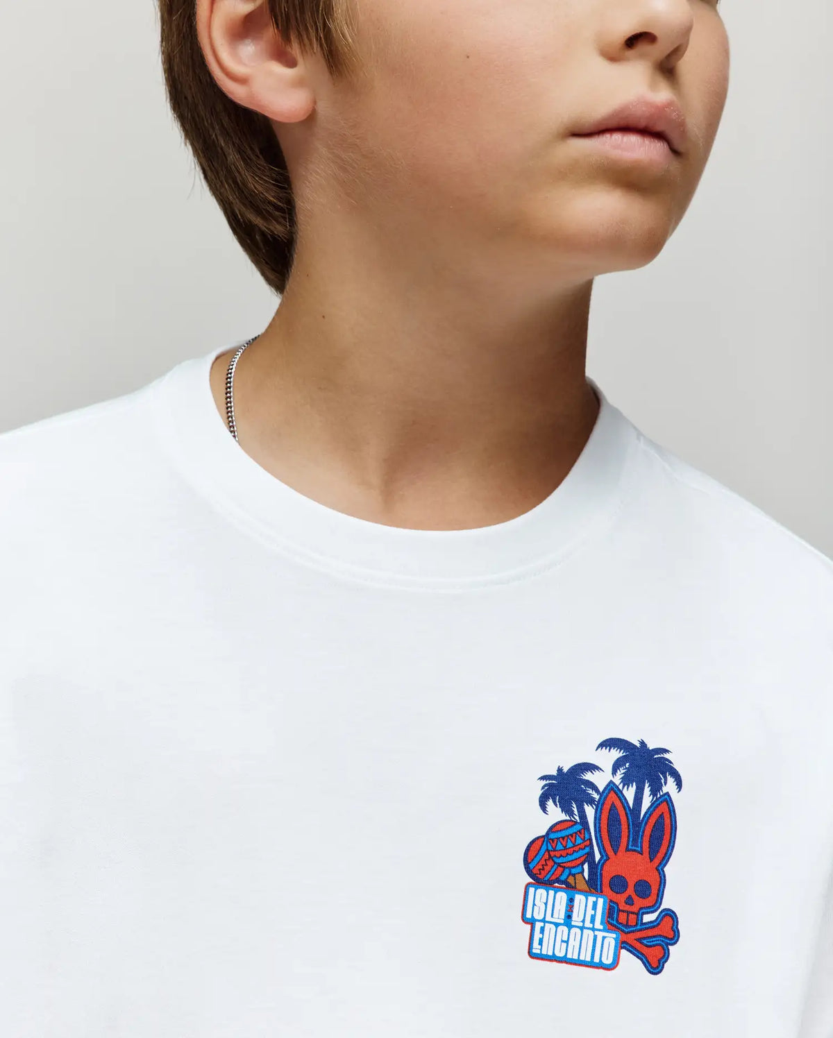 A young person wears the Psycho Bunny KIDS PUERTO RICO TEE (KT0200133), a white Pima cotton shirt with a vibrant bunny graphic, palm trees, and "Isla del Encanto." The face is partially out of frame.