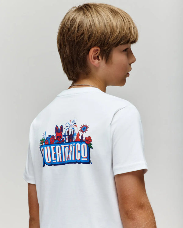 A young boy with light brown hair wears the Psycho Bunny KIDS PUERTO RICO TEE (KT0200133), a white Pima cotton shirt with colorful Puerto Rico-themed graphics and back text, standing against a plain light background.