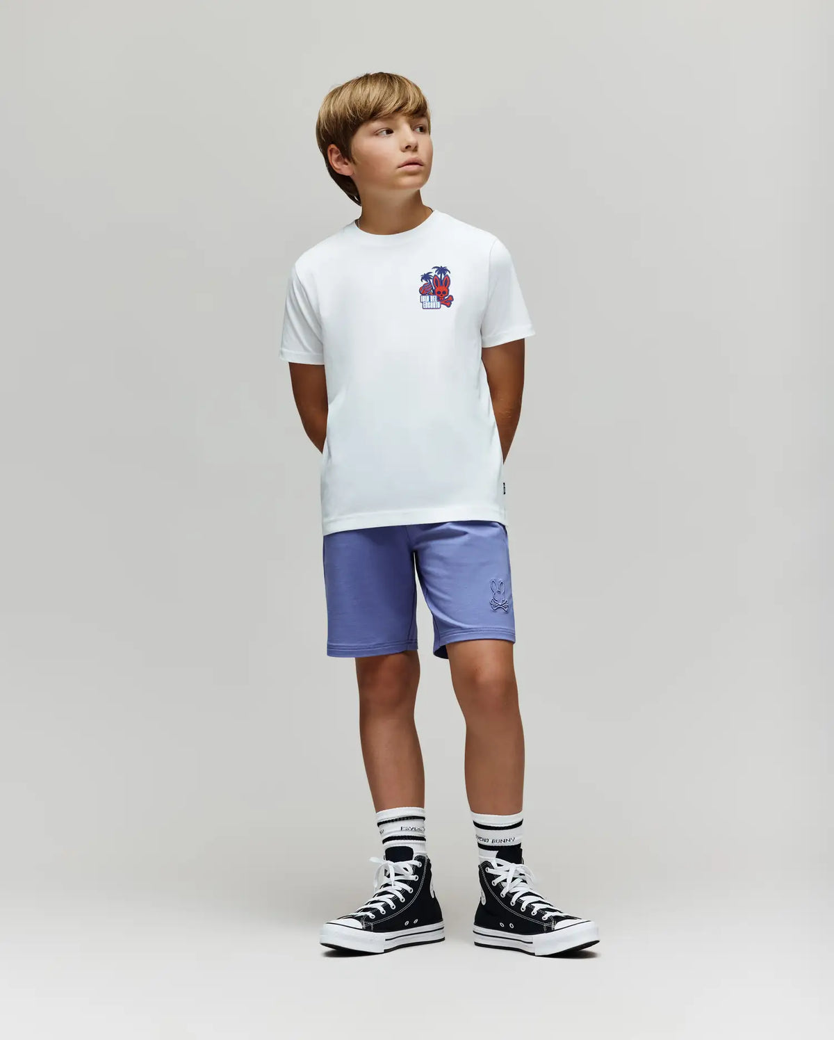 A young boy stands with hands behind his back, wearing the Psycho Bunny KIDS PUERTO RICO TEE (KT0200133), light purple shorts, striped socks, and black high-top sneakers against a plain light gray background.