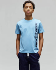 Kids Toby Wordmark Tee, a light blue classic fit jersey knit t-shirt featuring a large vertical dark blue Psycho Bunny wordmark and bunny skull logo print on the right side.