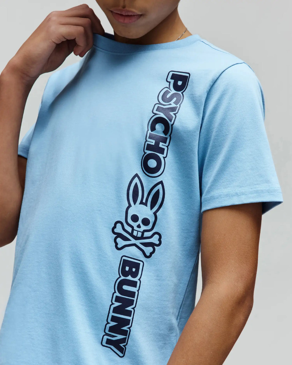 Kids Toby Wordmark Tee, a light blue jersey knit t-shirt with a classic fit, featuring a large vertical dark blue Psycho Bunny wordmark and iconic bunny skull logo graphic on the front.