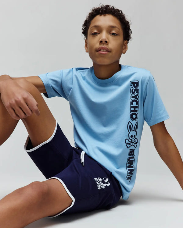 Kids Toby Wordmark Tee in light blue jersey, classic fit, with a large black vertical Psycho Bunny wordmark and bunny skull logo. Navy blue shorts with white trim and a small white bunny skull logo.