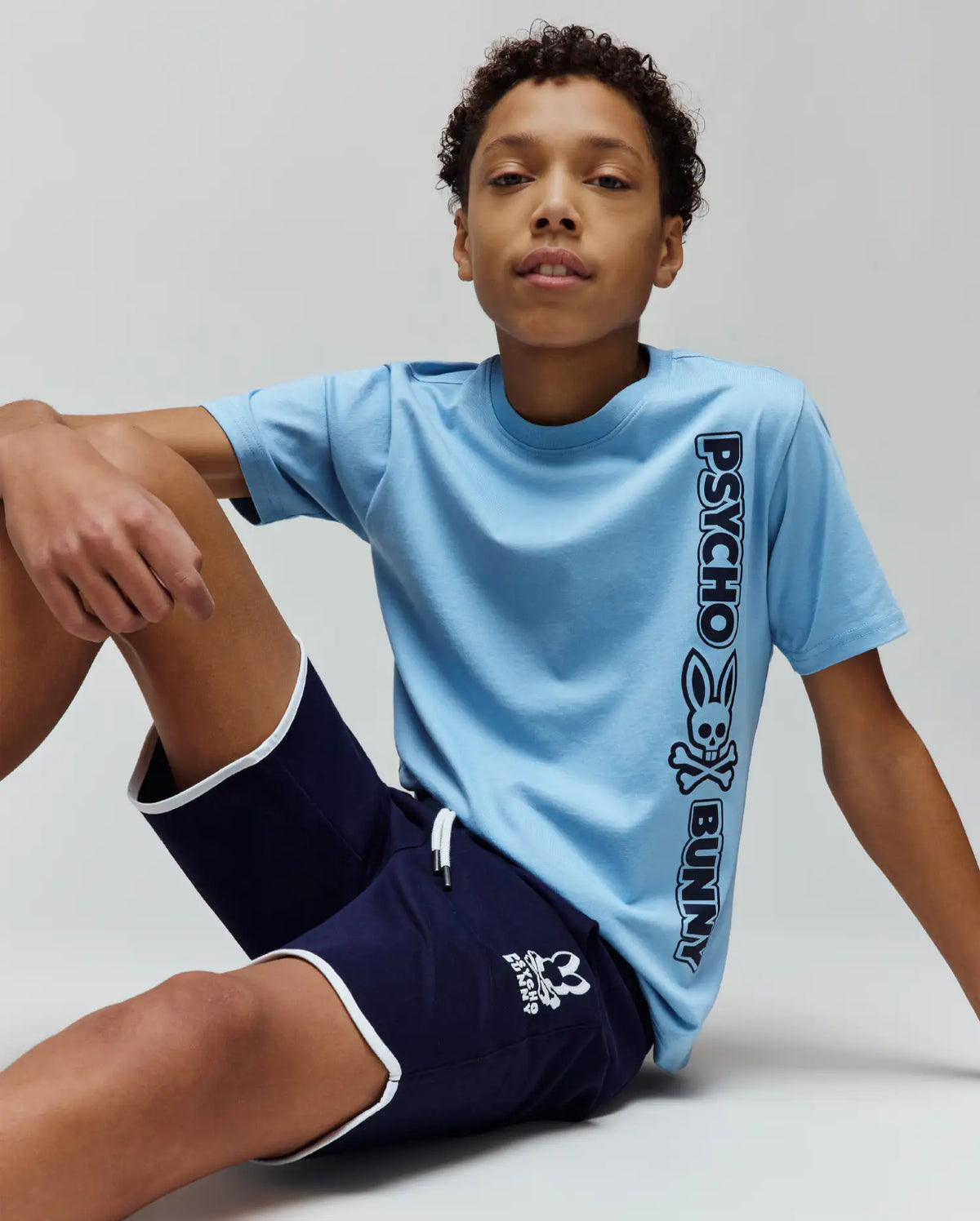 Kids Toby Wordmark Tee in light blue jersey, classic fit, with a large black vertical Psycho Bunny wordmark and bunny skull logo. Navy blue shorts with white trim and a small white bunny skull logo.