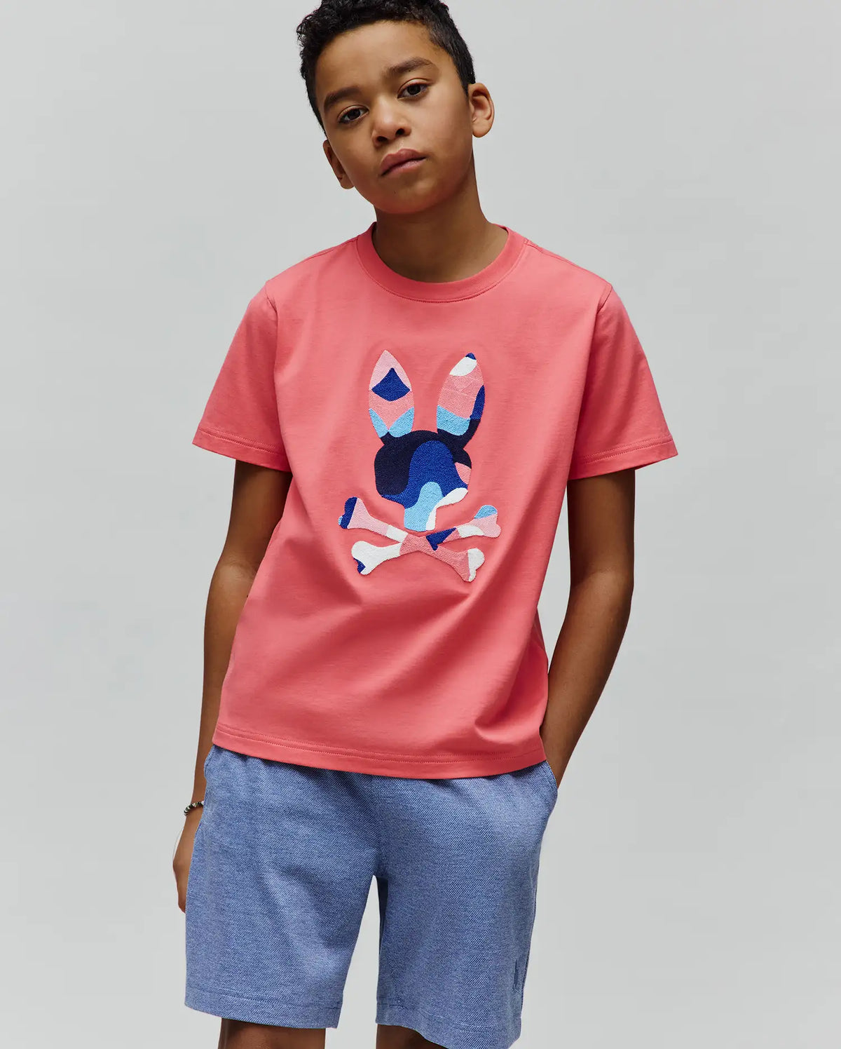 Kids Ara Graphic Tee, a coral classic fit jersey t-shirt featuring a large, multi-toned blue, pink, and white patterned Psycho Bunny skull logo on the center chest.