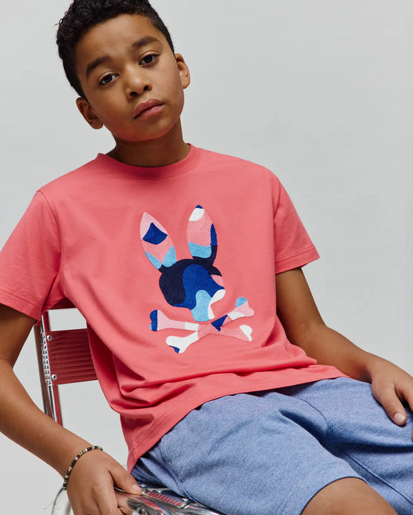 Kids Ara Graphic Tee in coral pink, featuring a large, abstract blue, pink, and white camo-style Psycho Bunny skull graphic on the chest, classic fit jersey knit.