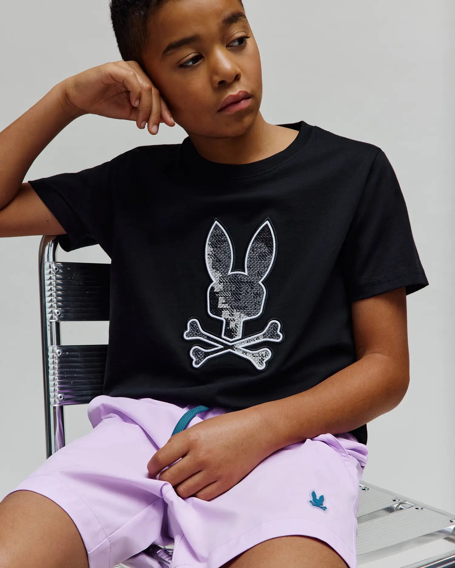 Kids Olivier Graphic Tee in black jersey, featuring a large silver sequin Psycho Bunny skull and crossbones chest graphic, paired with lavender woven shorts displaying a small embroidered teal bunny logo.