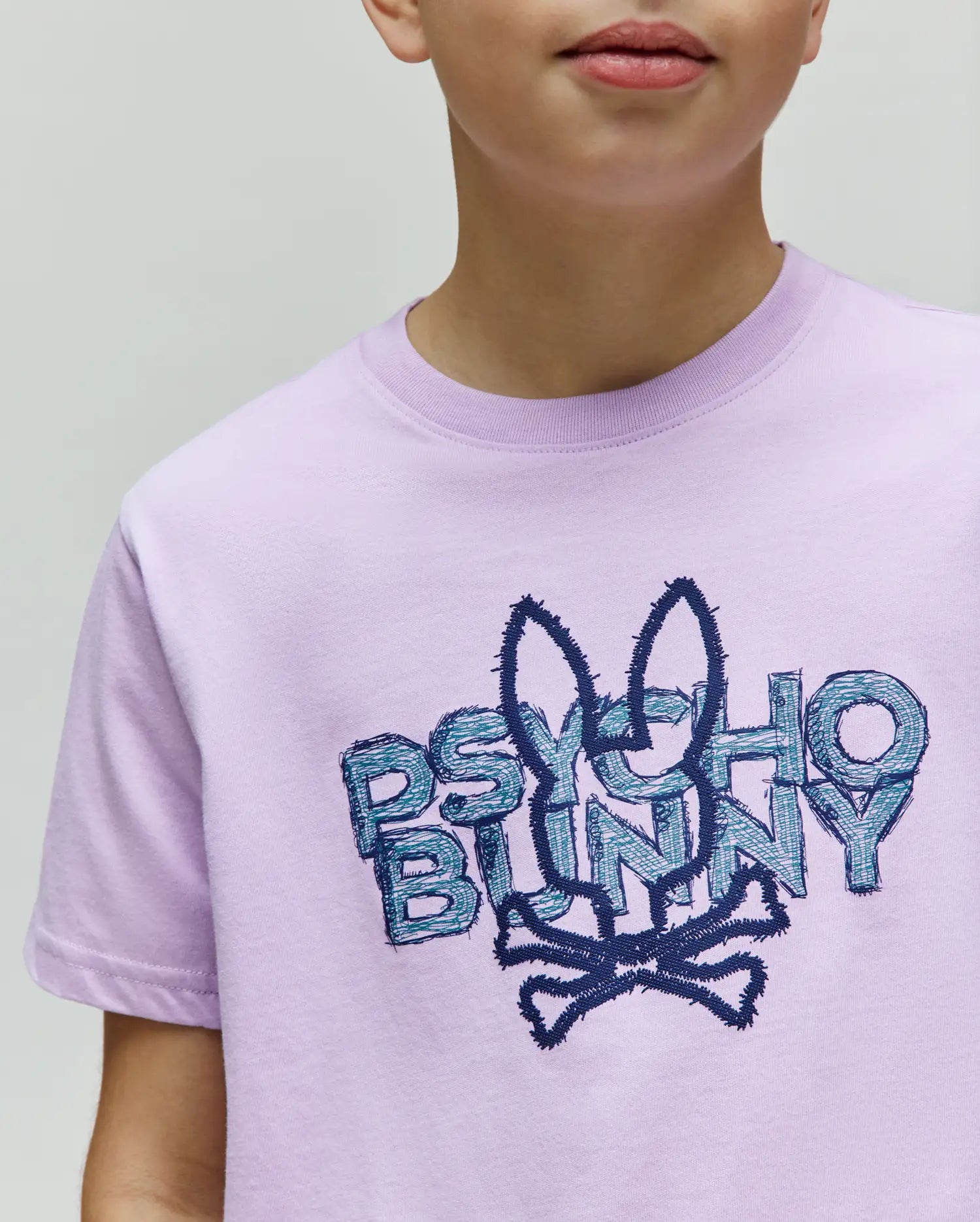 Kids Herman Graphic Tee, a lavender classic fit jersey knit t-shirt featuring a large, textured dark blue and teal Psycho Bunny logo with skull graphic on the central chest.