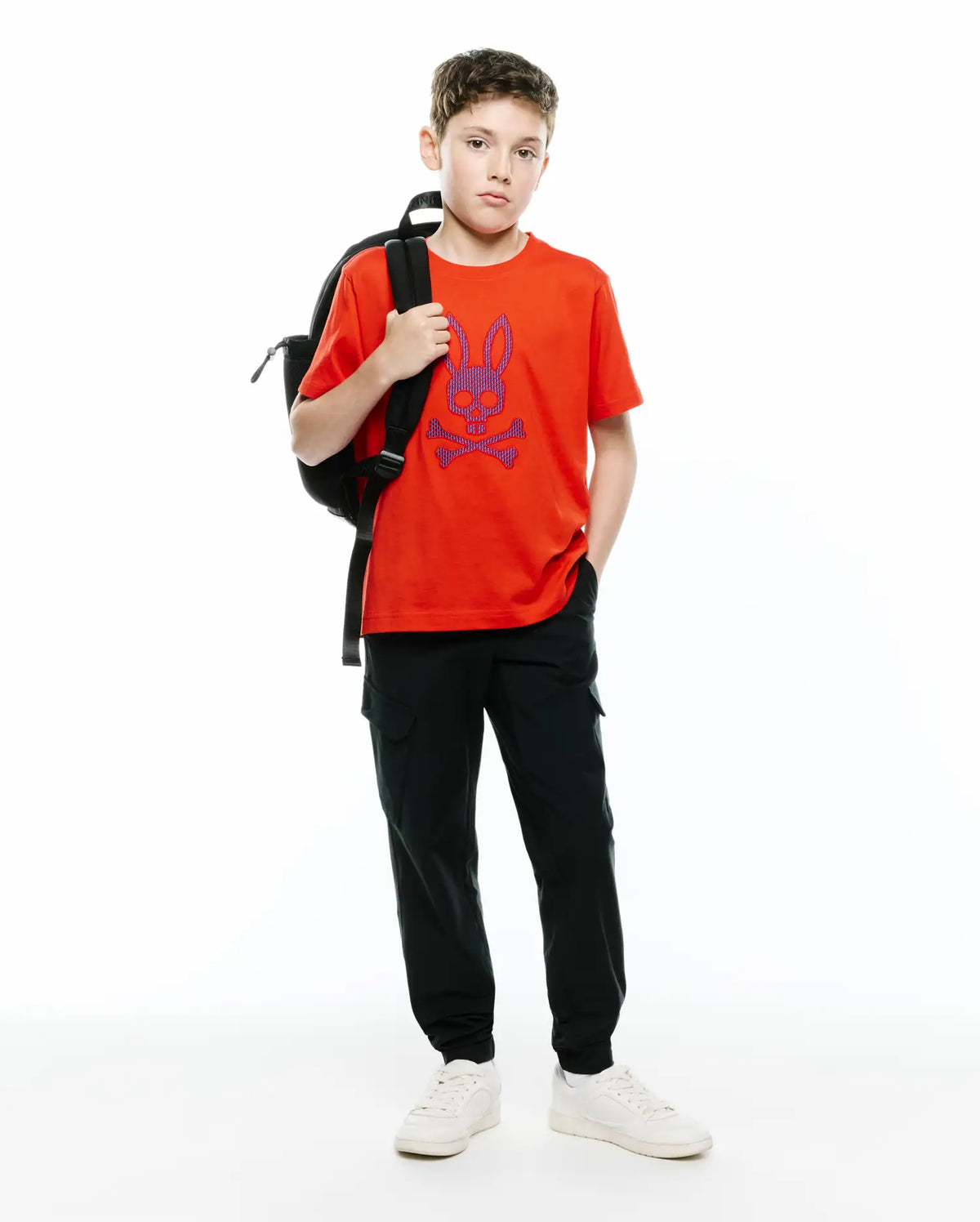 Vibrant orange Kids Stan Graphic Tee, a classic fit jersey t-shirt featuring a large purple and blue Psycho Bunny skull logo on the chest, paired with relaxed dark green cargo pants. (5)