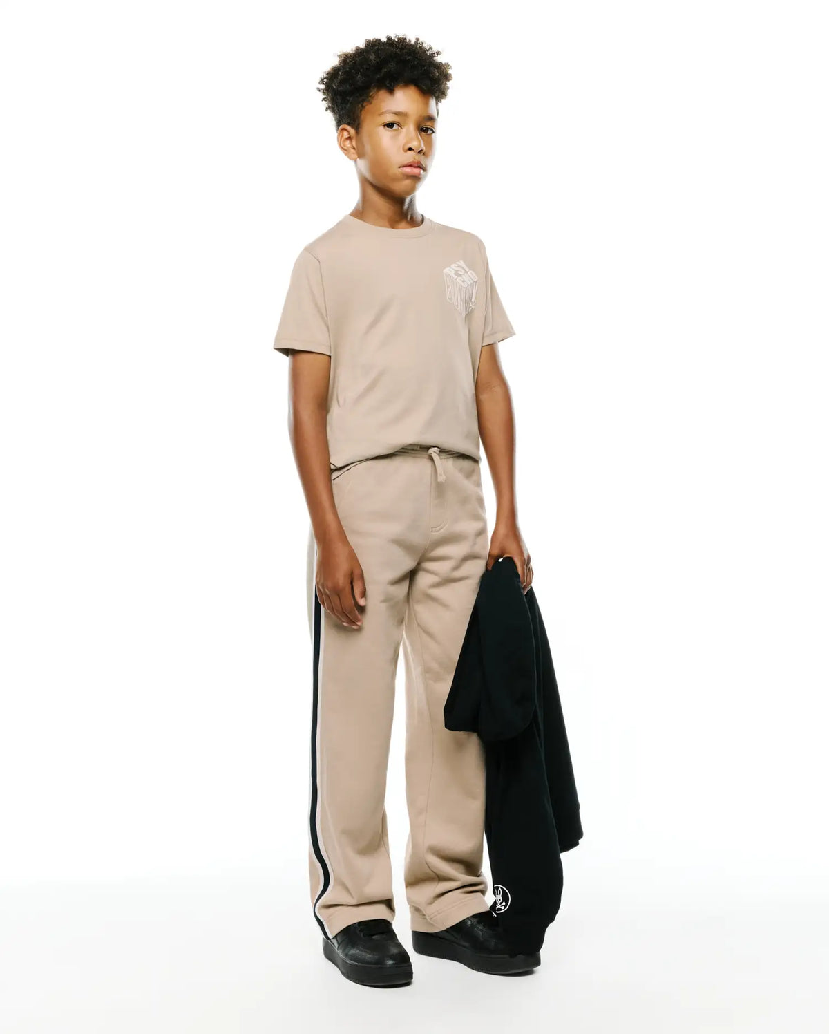 Beige jersey KIDS CONAN BACK GRAPHIC TEE with a white dripping Psycho Bunny chest graphic, matching relaxed fleece sweatpants with black and white side stripes, and a dark hoodie with a sleeve logo. (4)