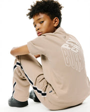 Kids Conan Back Graphic Tee, beige jersey with a large white 3D Psycho Bunny graphic on back, and relaxed beige track pants featuring black/white side stripes and an embroidered bunny skull. (1)