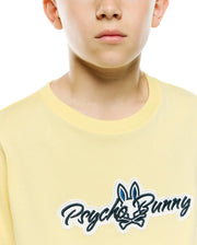 Kids Jaydon Wordmark Graphic Tee in light yellow jersey knit, featuring a large embroidered Psycho Bunny wordmark logo on the chest with a dark blue bunny skull and white outline. (3)