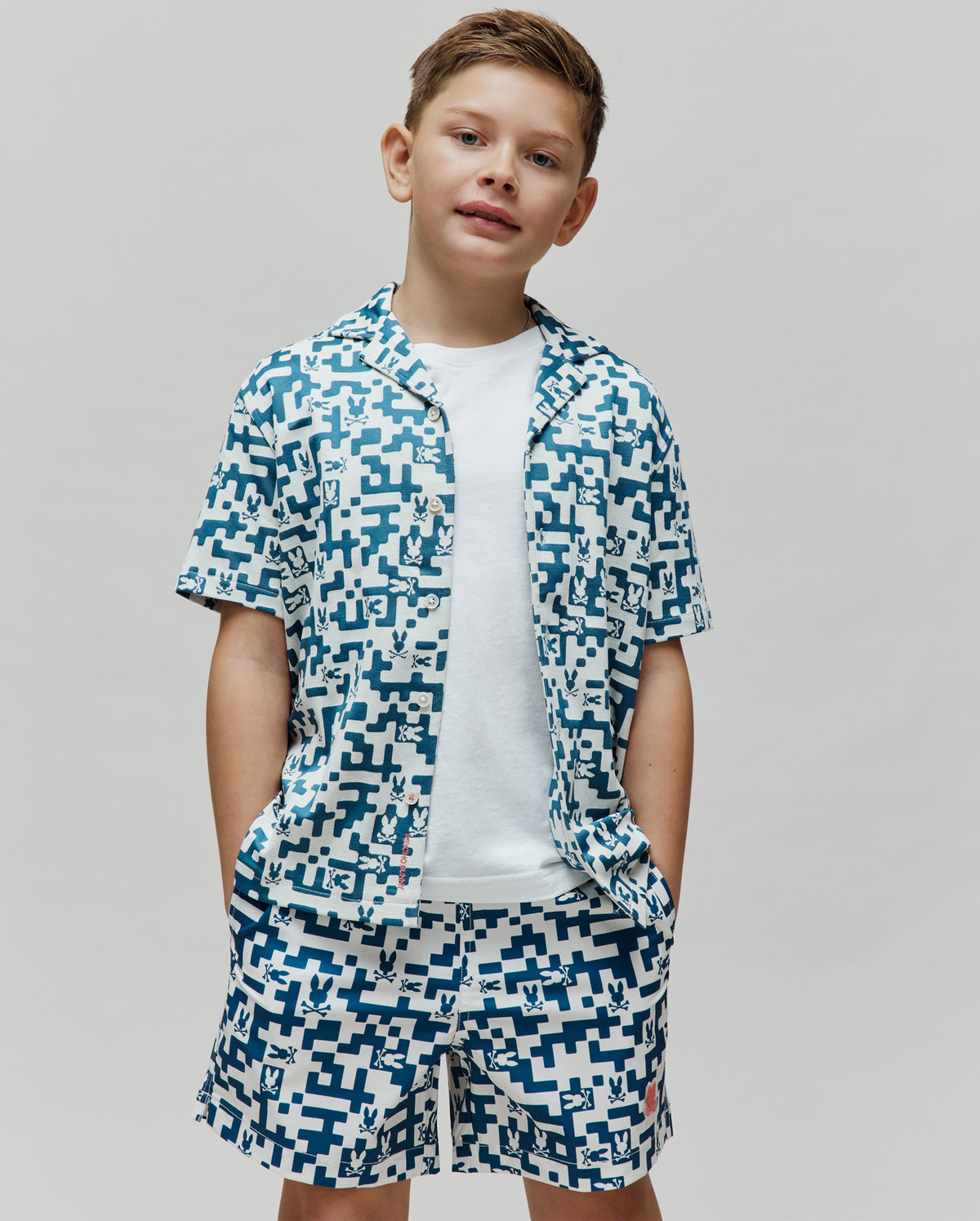 Kids Josh Jacquard Shirt and matching shorts, a relaxed fit white set with an all-over teal pixelated geometric pattern featuring small Psycho Bunny skull logos woven into the jacquard fabric.