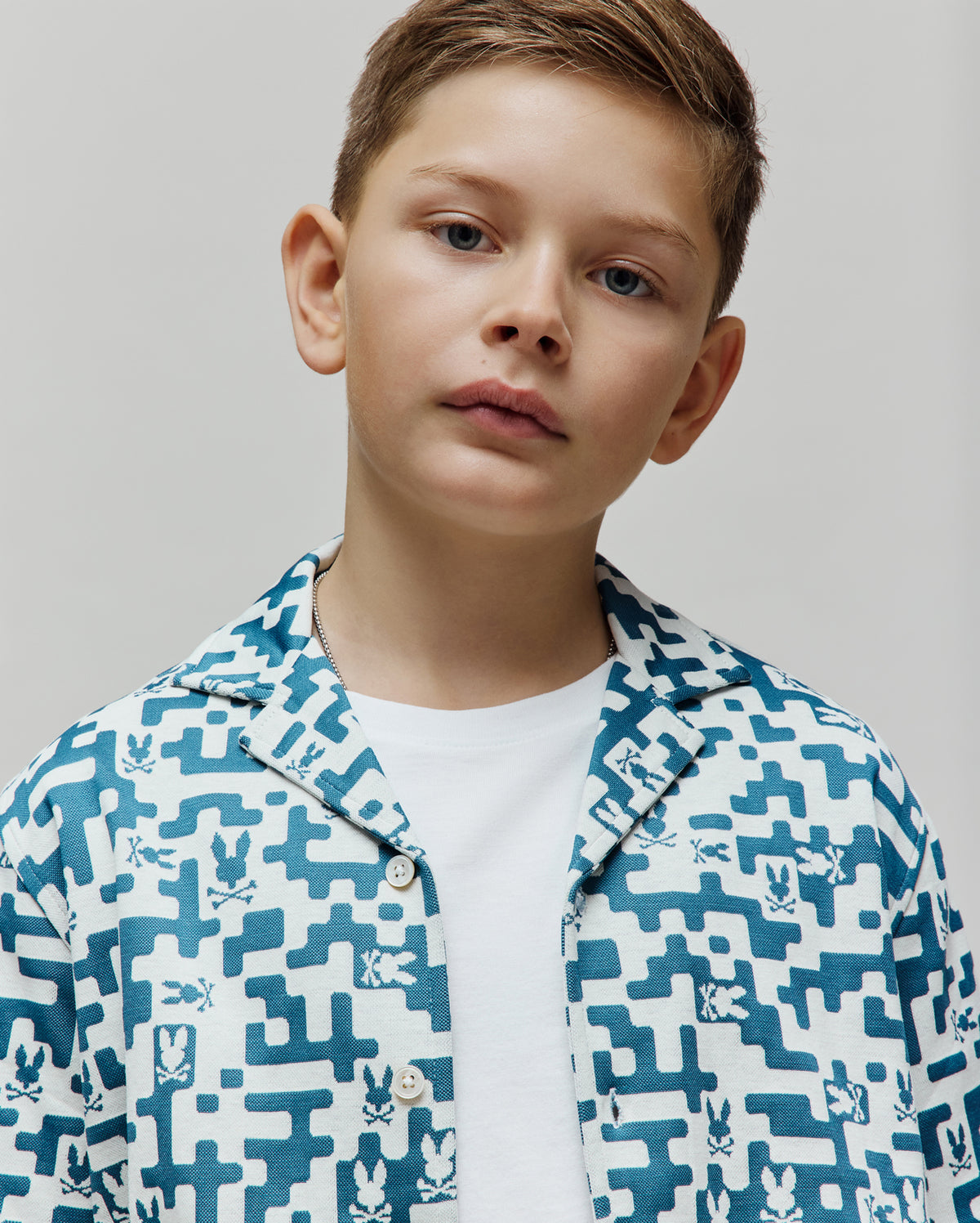 Kids Josh Jacquard Shirt, a white and teal blue jacquard knit short-sleeve shirt with an all-over geometric pattern featuring small Psycho Bunny skull logos, classic fit.