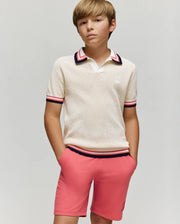 A boy with light brown hair wears the Psycho Bunny KIDS CARTER KNIT POLO SHIRT (KT0400006), a cream, short-sleeved, 100% cotton polo with Johnny collar and red, navy, and white trim, standing hands-in-pockets against a gray background.