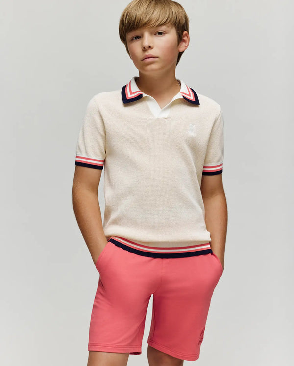 A boy with light brown hair wears the Psycho Bunny KIDS CARTER KNIT POLO SHIRT (KT0400006), a cream, short-sleeved, 100% cotton polo with Johnny collar and red, navy, and white trim, standing hands-in-pockets against a gray background.