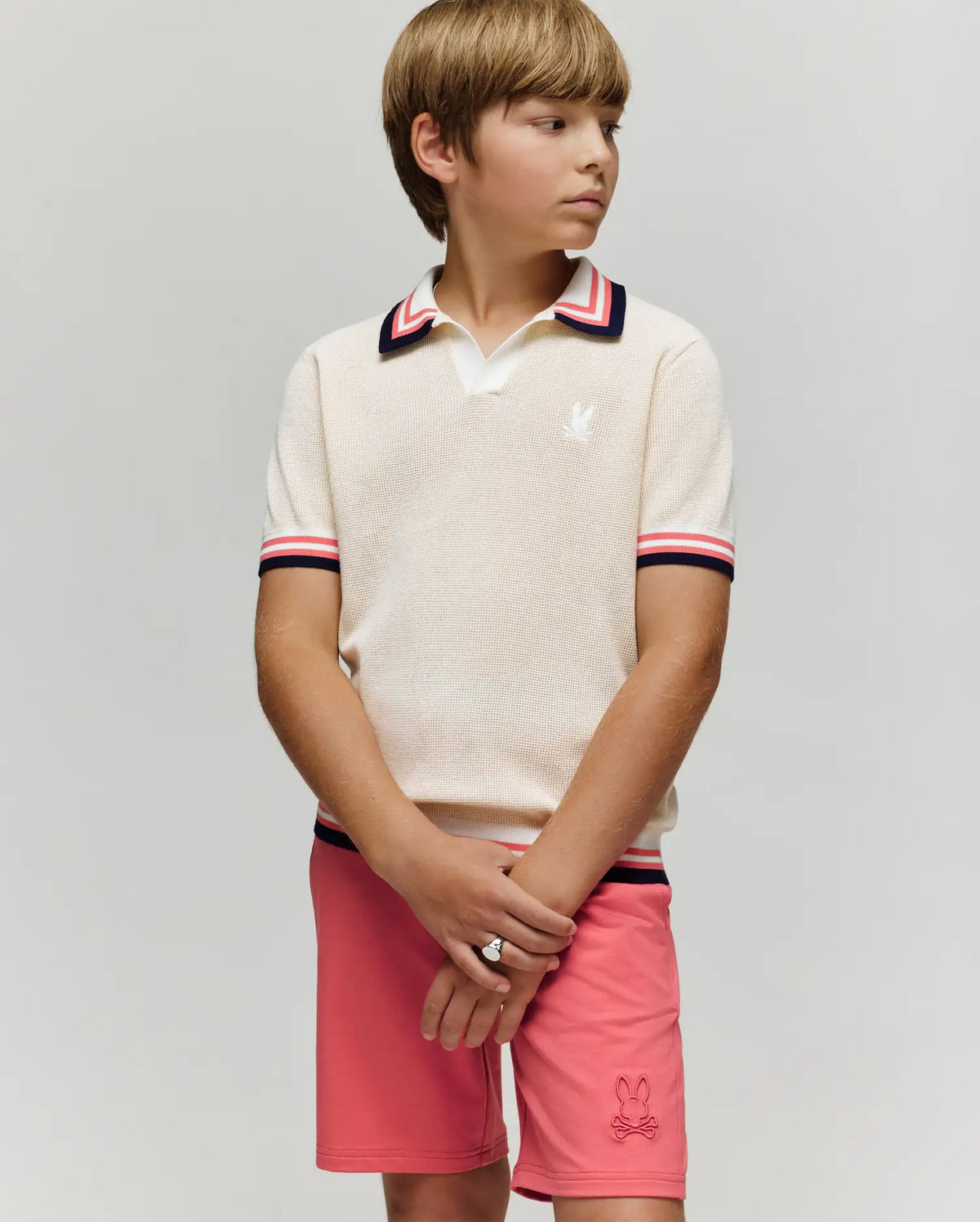 A young boy stands against a plain background, wearing the Psycho Bunny KIDS CARTER KNIT POLO SHIRT - KT0400006 in cream with red, navy, and white trim, paired with pink shorts featuring a small bunny logo. He looks to the side, hands clasped.