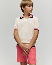 A young boy stands against a plain background, wearing the Psycho Bunny KIDS CARTER KNIT POLO SHIRT - KT0400006 in cream with red, navy, and white trim, paired with pink shorts featuring a small bunny logo. He looks to the side, hands clasped.