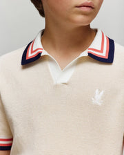 A young person wears the KIDS CARTER KNIT POLO SHIRT (KT0400006) by Psycho Bunny—cream, 100% cotton, with a white, red, and navy Johnny collar and embroidered chest logo. Only their lower face and torso are visible.