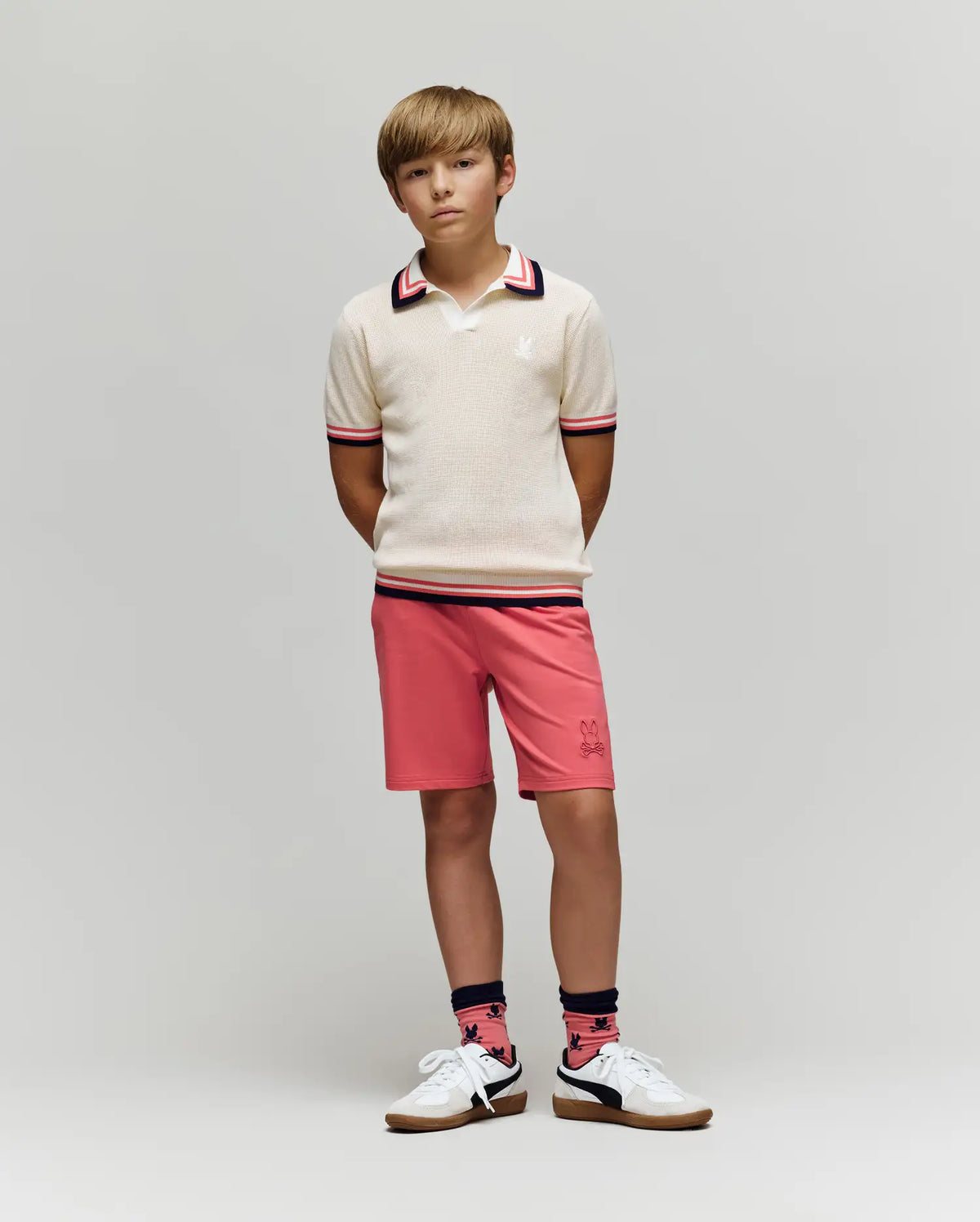 A boy stands against a plain background wearing the Psycho Bunny KIDS CARTER KNIT POLO SHIRT - KT0400006, paired with salmon pink shorts, pink and navy socks, and white sneakers. His hands are behind his back for a relaxed look.