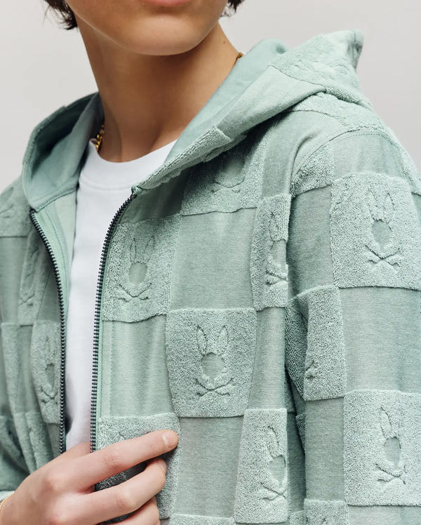 A person wears the Psycho Bunny KIDS KELVIN FULL ZIP TOWEL TERRY HOODIE (KT0500004), a light green cotton-blend hoodie with a checkerboard Bunny pattern and raised skulls, over a white t-shirt. Only their lower face and torso are visible.