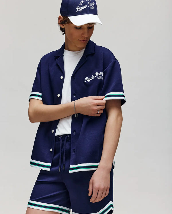 A child poses against a light background in Psycho Bunny’s KIDS BASIL TAPED CAMP COLLAR SHIRT (KT0500007), paired with a matching navy cap and shorts, all with embroidered logos and white-green striped trim, crafted from a premium cotton-modal blend.