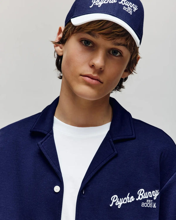 A young person wears the Psycho Bunny KIDS BASIL TAPED CAMP COLLAR SHIRT (KT0500007), a navy blue piece with the embroidered "Psycho Bunny EST. 2005" logo, paired with a matching cap and white cotton-modal tee against a neutral background.