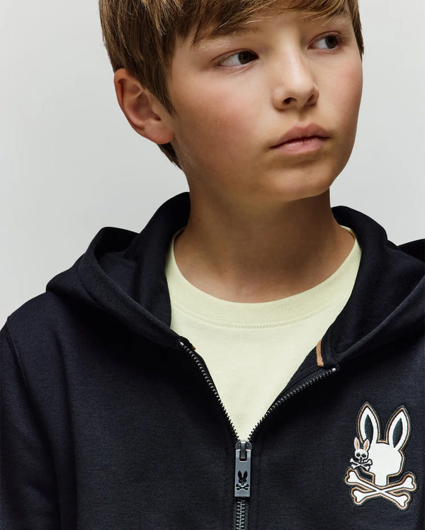 A young boy wears the Psycho Bunny KIDS WADE CORD PATCH ZIP HOODIE (KT0500016), featuring an embroidered Bunny and crossbones logo, over a light T-shirt, gazing off to the side against a plain background.