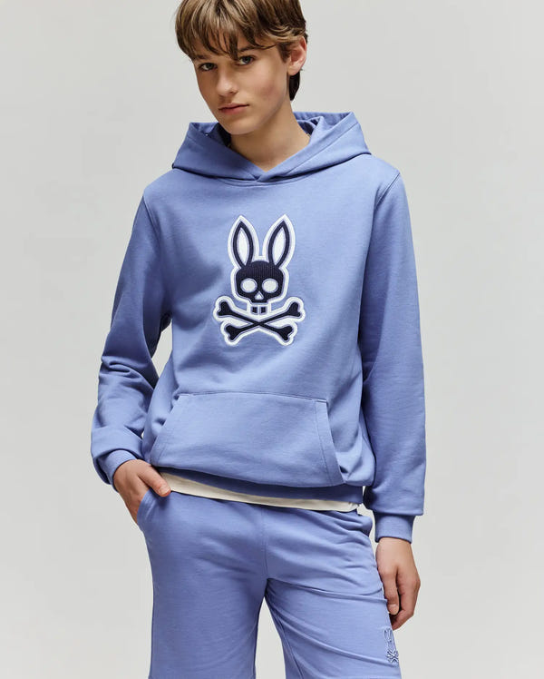 A young person wears the Psycho Bunny KIDS RYDER BUNNY HOODIE - KT0500018 in light blue, featuring a bold, embroidered Bunny skull and crossbones patch—perfect for stylish loungewear.