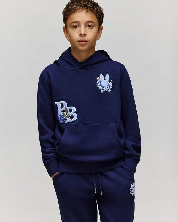 A boy with short brown hair wears the Psycho Bunny KIDS AUGUSTE HOODIE (KT0500019), a navy blue cotton fleece hoodie with matching sweatpants, both featuring embroidered designs like a rabbit and "PB." He stands hands in pockets against a plain background.