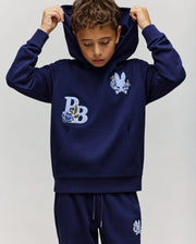 A young boy wears the Psycho Bunny KIDS AUGUSTE HOODIE (KT0500019), a regular fit cotton fleece with bunny and "PB" patches, pulling the hood over his head. He pairs it with matching navy blue sweatpants.