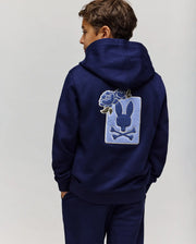 A boy faces away from the camera wearing the Psycho Bunny KIDS AUGUSTE HOODIE (KT0500019) in navy blue, featuring a cotton fleece regular fit and a large bunny silhouette with crossbones and chenille floral patch on the back.