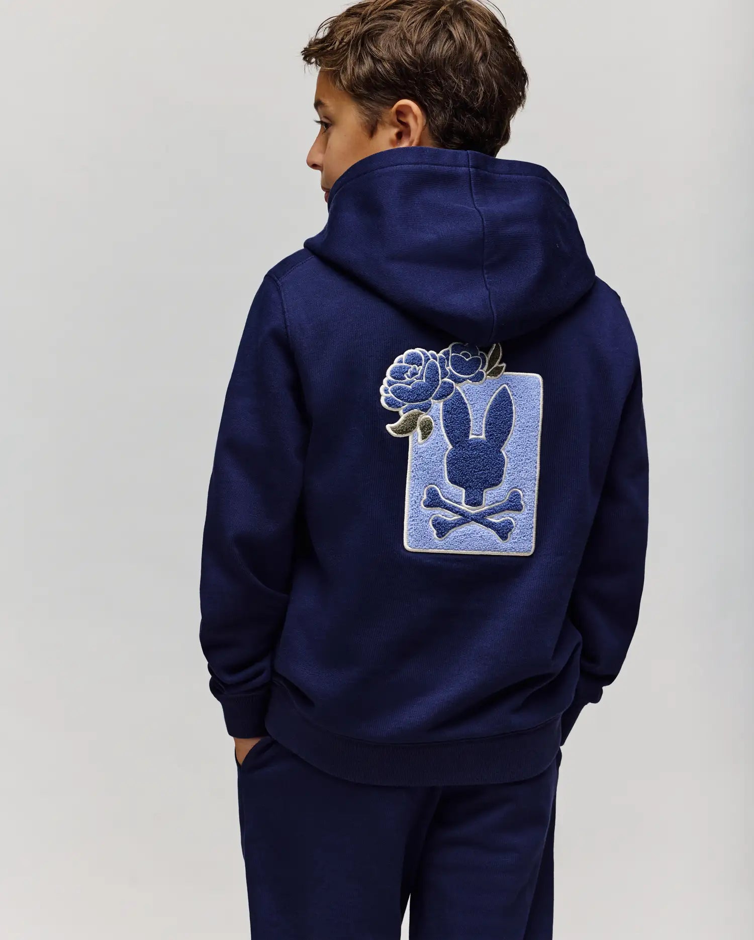 A boy faces away from the camera wearing the Psycho Bunny KIDS AUGUSTE HOODIE (KT0500019) in navy blue, featuring a cotton fleece regular fit and a large bunny silhouette with crossbones and chenille floral patch on the back.
