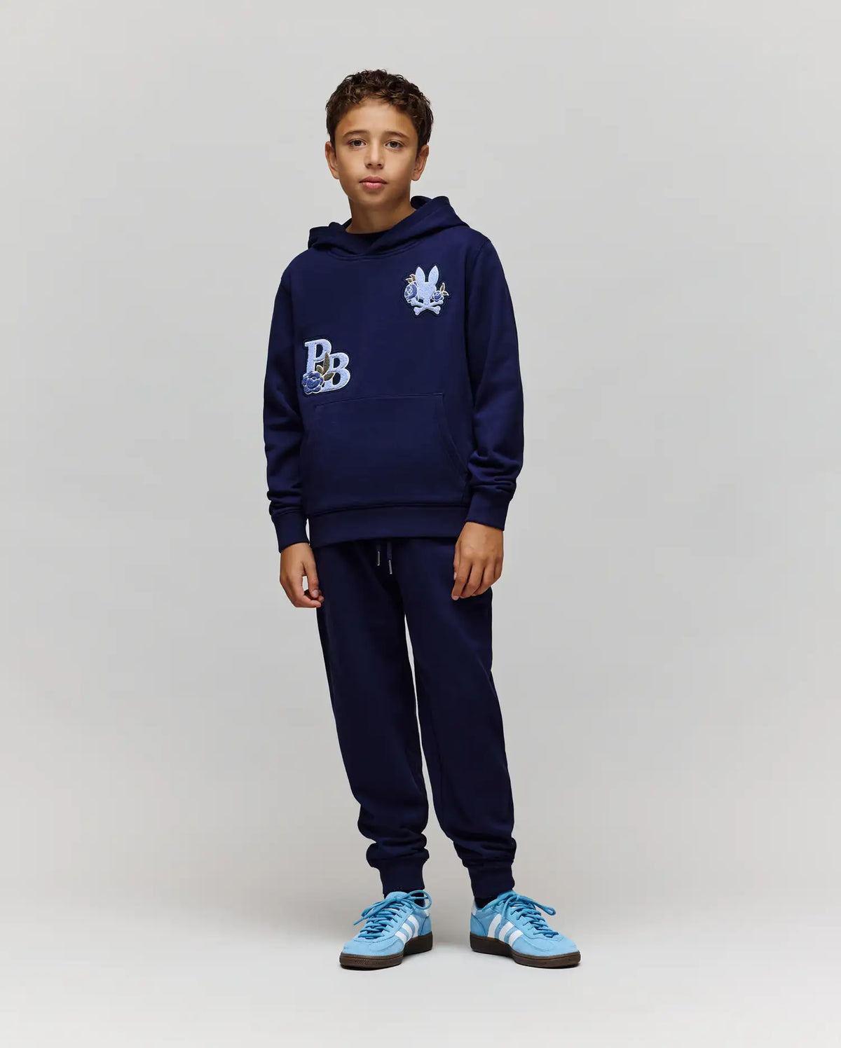 A boy faces forward wearing the Psycho Bunny KIDS AUGUSTE HOODIE - KT0500019 in navy blue with graphic patches and chenille floral details, paired with matching navy sweatpants and blue sneakers, set against a plain light gray background.