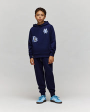A boy faces forward wearing the Psycho Bunny KIDS AUGUSTE HOODIE - KT0500019 in navy blue with graphic patches and chenille floral details, paired with matching navy sweatpants and blue sneakers, set against a plain light gray background.