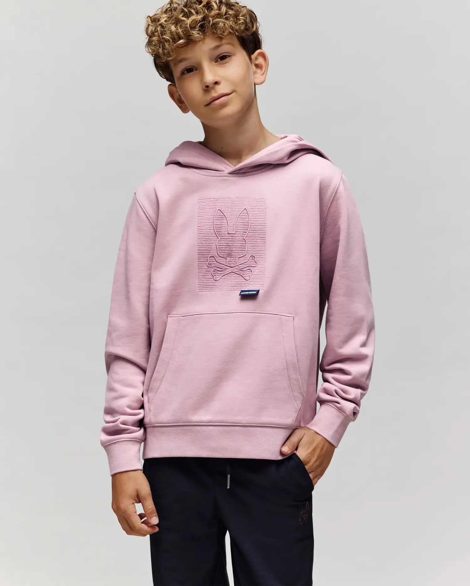 A curly-haired boy stands against a plain light gray background, wearing the Psycho Bunny KIDS GRANT HOODIE (KT0500020) in light pink, featuring the brand's bunny and crossbones design on the front.