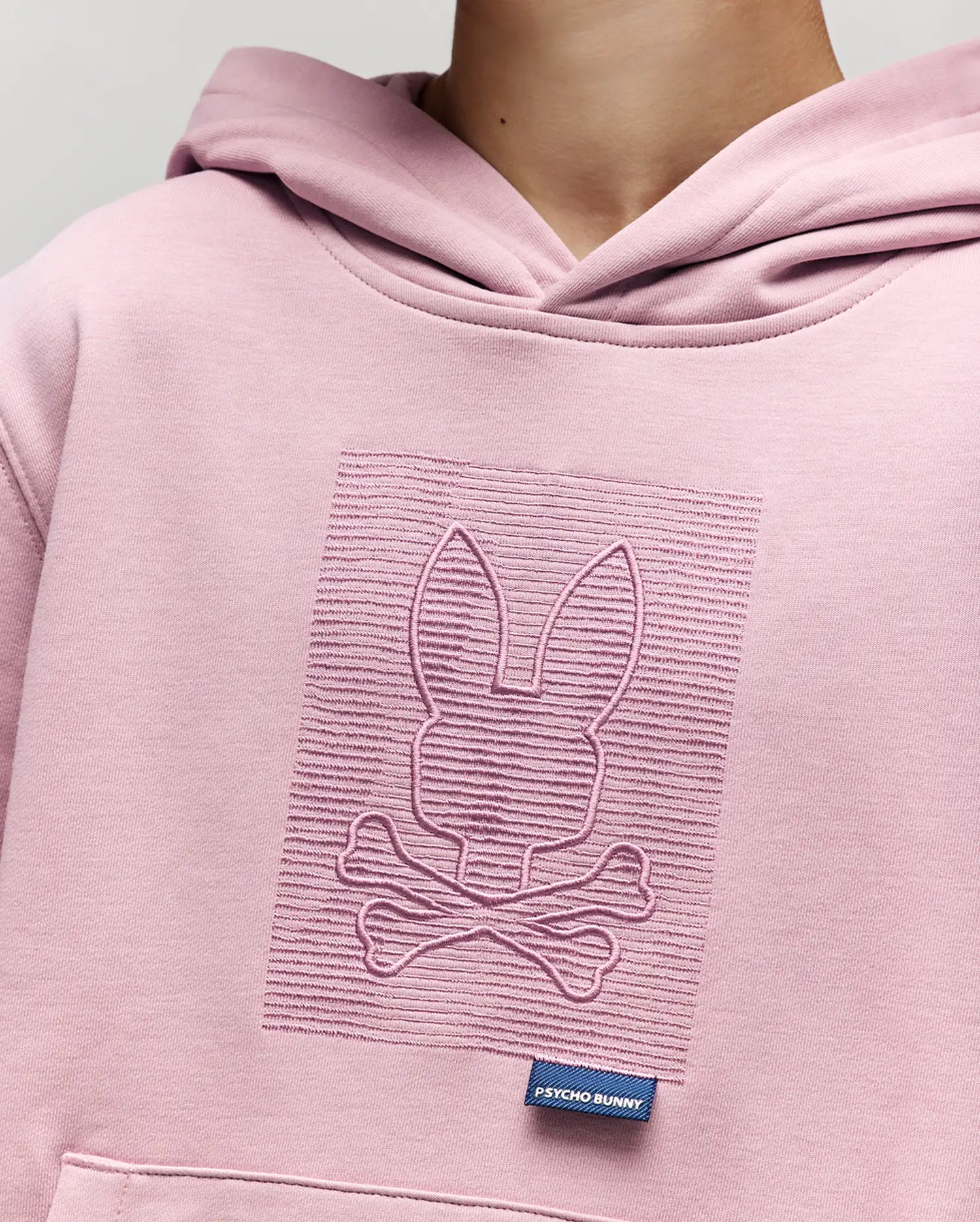 A close-up of a child wearing the Psycho Bunny KIDS GRANT HOODIE in light pink, showcasing an embossed bunny and crossbones design on the chest and a small blue Psycho Bunny label on the pocket.