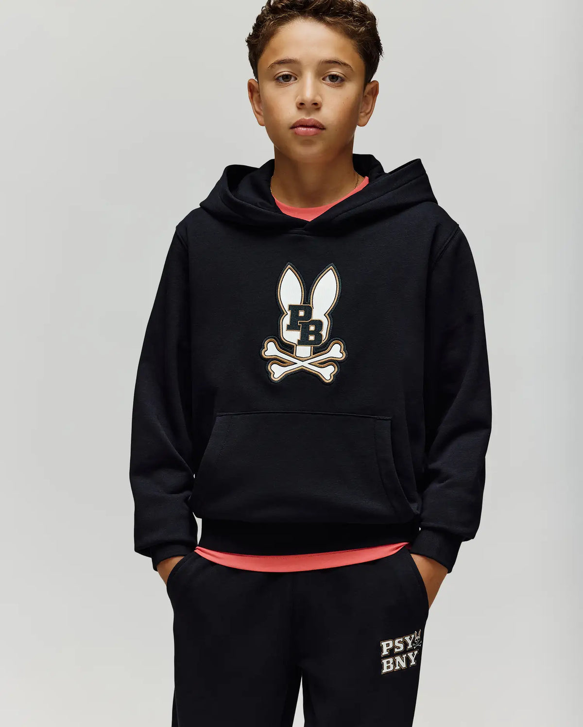 A young boy with short curly hair wears the Psycho Bunny KIDS WADE CORD PATCH HOODIE (KT0500031) and matching black pants with "PSYCHO BNY" on the pocket, standing against a plain light background.