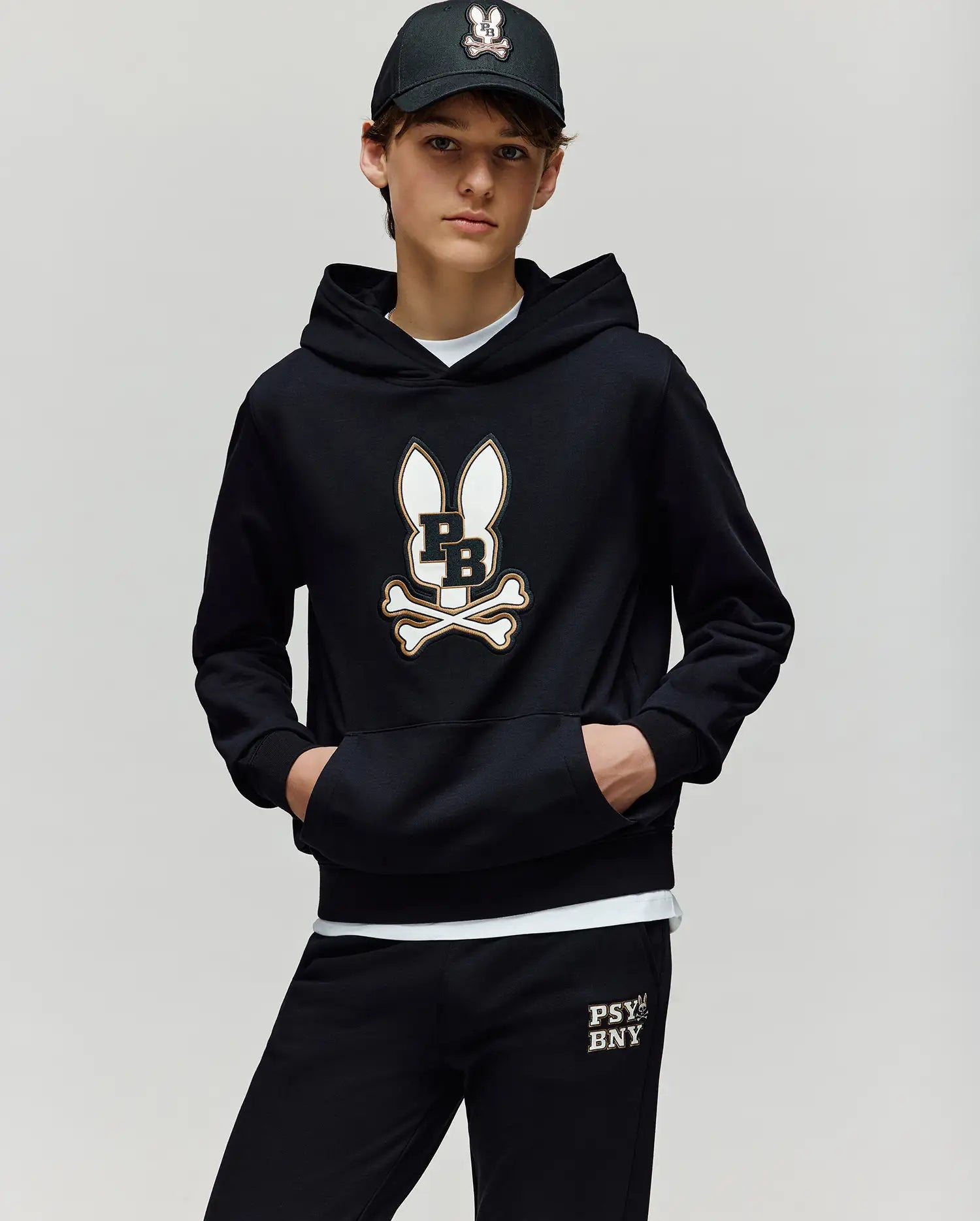 A boy wears the Psycho Bunny KIDS WADE CORD PATCH HOODIE (KT0500031) and matching pants, both in premium cotton blend with white and gold bunny and crossbones logos. His black cap features an embroidered Bunny patch, standing against a plain background.